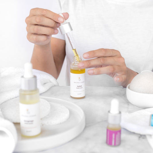 Original Human Co. Facial Serums Putting Your Skin Care Routine In Order - Image of someone sitting at a white table with an open bottle of Original Human Co. Facial Serum in front of them amongst other closed bottles. 